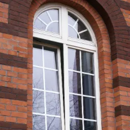 Otwarta okno z białymi szprosami w łukowatej, ceglanej wnęce, odbicie drzewa w szybie.