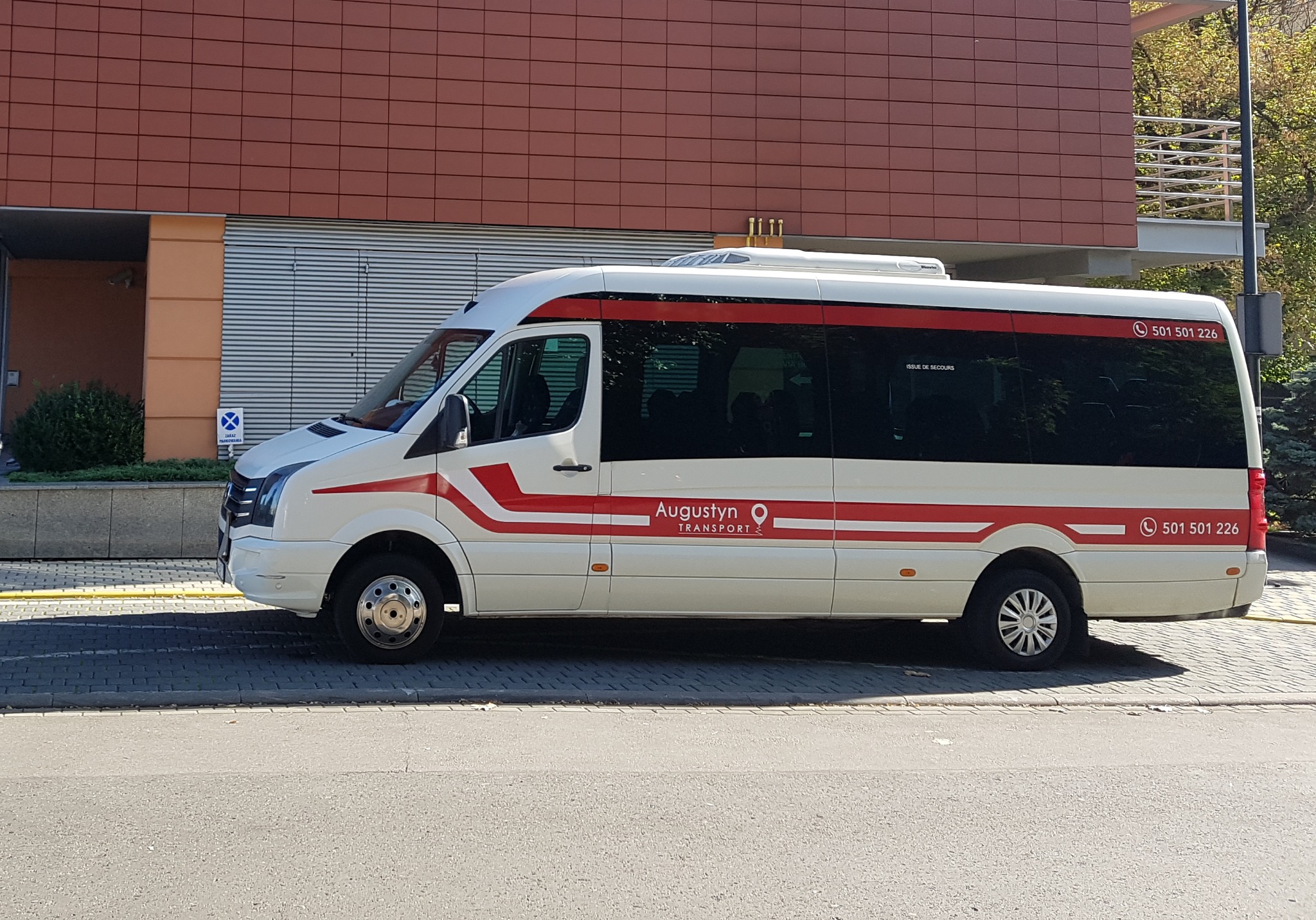 Biały bus z czerwonymi pasami i logo firmy Augustyn Transport zaparkowany na ulicy, widok z boku na tle budynku z brązową elewacją.