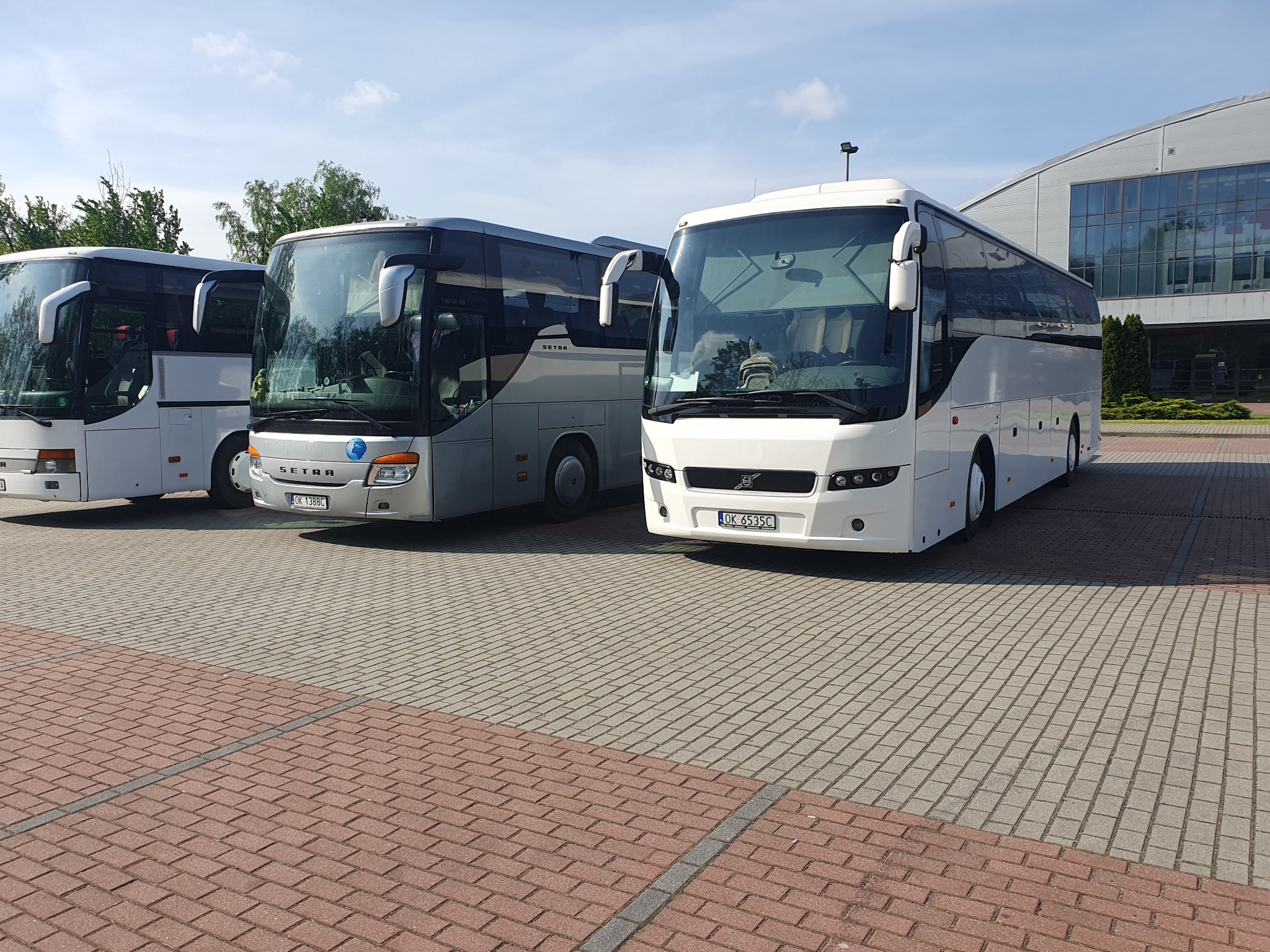 Trzy autokary Setra zaparkowane na placu z kostki, biały i srebrny, widok z przodu, budynek w tle.