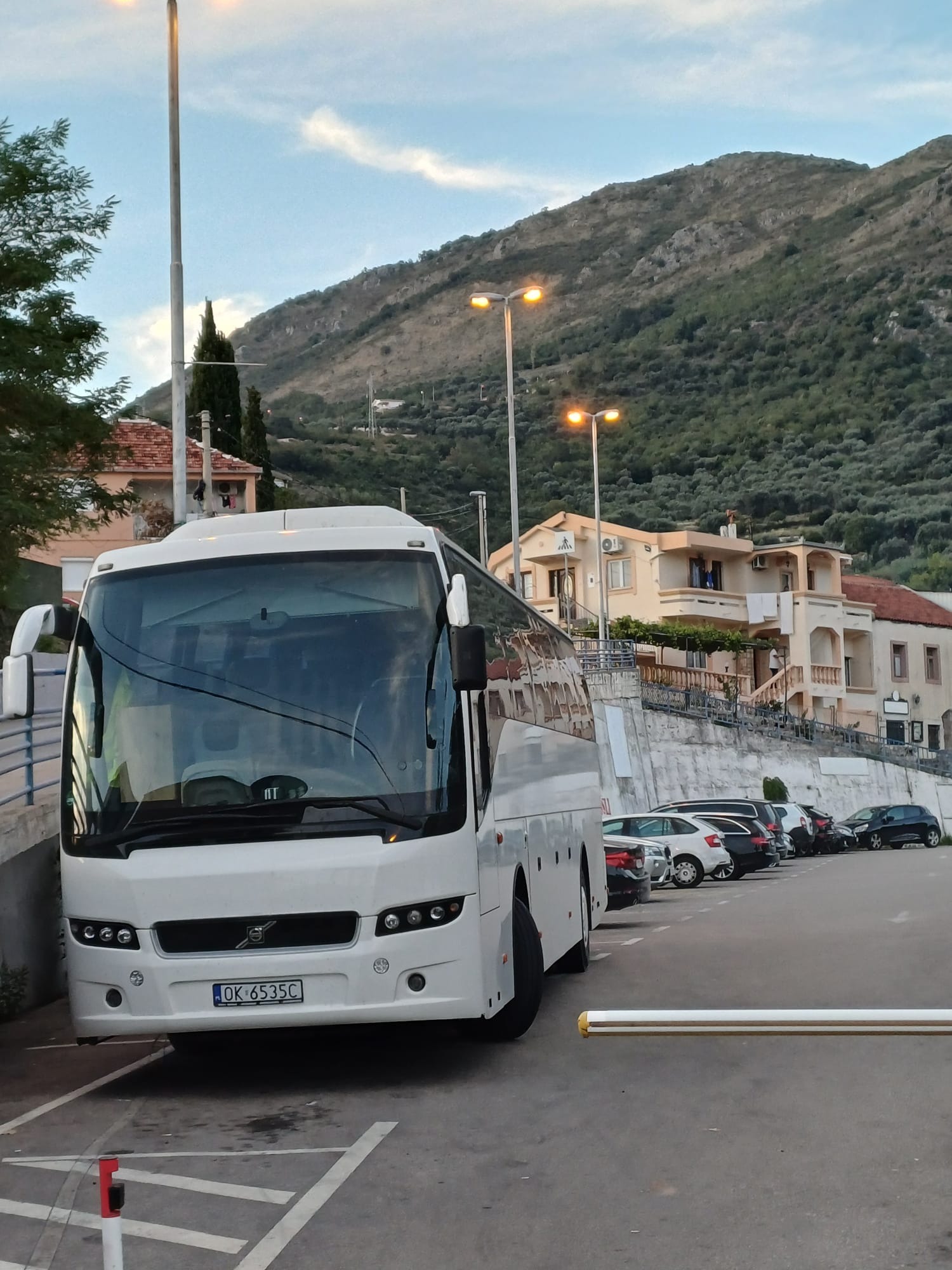 Biały autokar Volvo zaparkowany na parkingu z widokiem na górzysty krajobraz i zabudowania mieszkalne w tle.