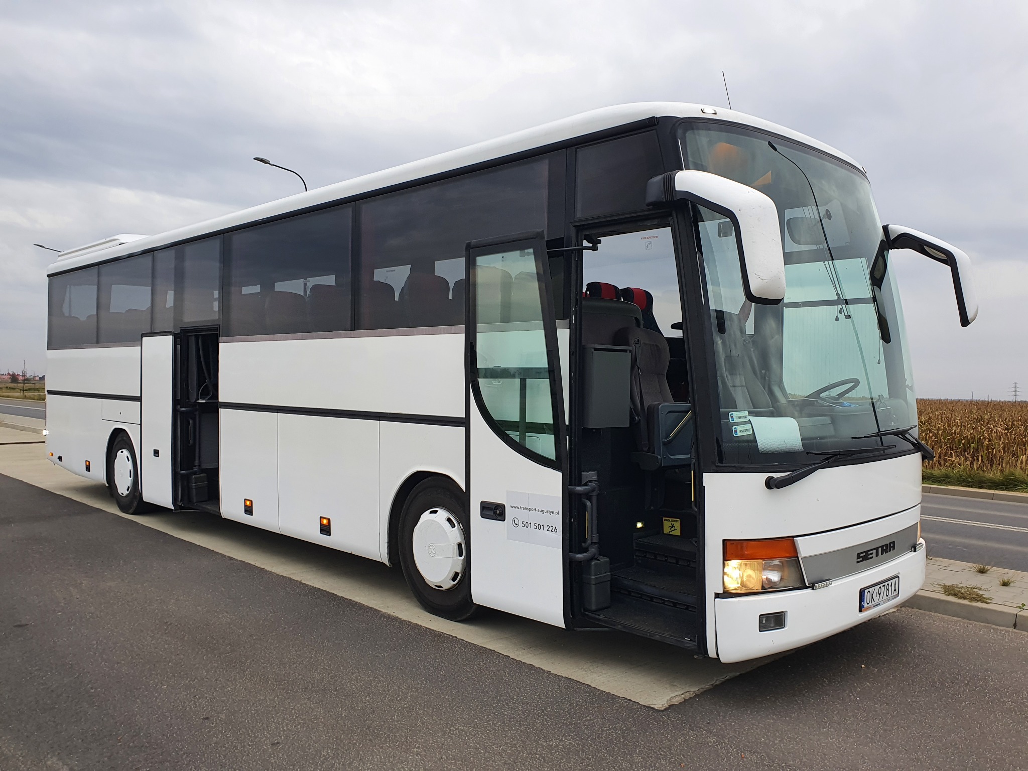 Biały autokar Setra z otwartymi drzwiami, zaparkowany na poboczu drogi, z widocznym numerem telefonu kontaktowego na boku pojazdu.