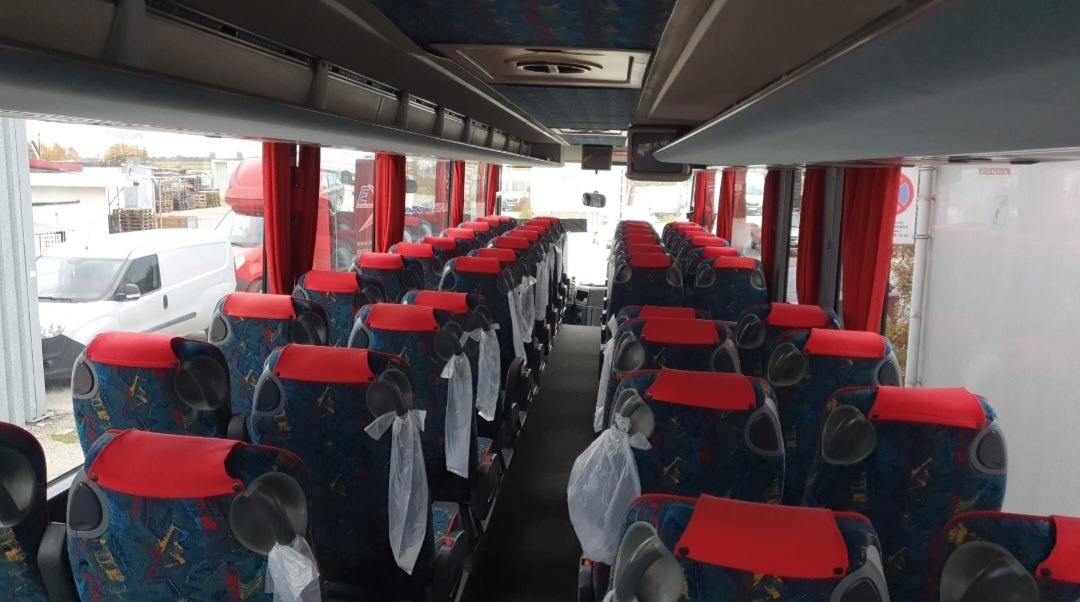 Wnętrze autobusu z rzędami foteli z czerwonymi zagłówkami i wzorzystą tapicerką, niektóre z plastikowymi osłonami, widok na zewnątrz przez okna z czerwonymi zasłonami.