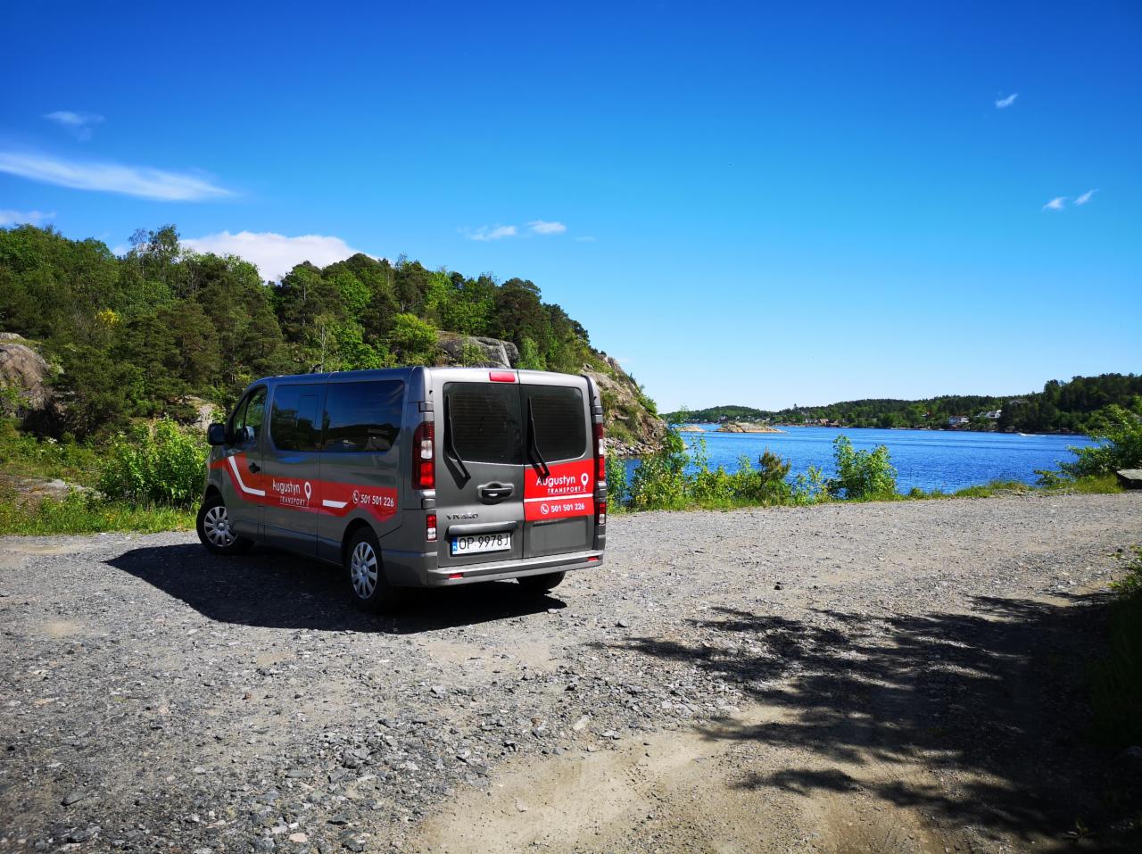 Szary bus firmy transportowej z logo na tle malowniczego krajobrazu z jeziorem i lasem w słoneczny dzień. Widoczne numery kontaktowe na pojeździe.