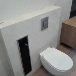 Podtynkowa toaleta wisząca z przyciskiem spłukującym i czarną wnęką rewizyjną, wykończona płytkami imitującymi jasny kamień, na drewnianej podłodze.