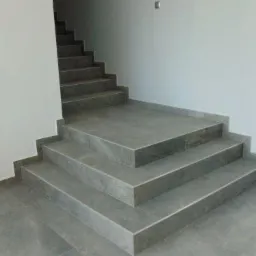 Wyłożone szarymi płytkami schody wewnątrz budynku, widoczna drabina i odkurzacz przemysłowy po lewej stronie, duże okno po prawej.