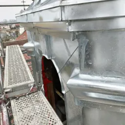 Szczegółowe ujęcie blaszanej obudowy wieży kościelnej z rusztowaniem, ukazujące detale łączeń i obróbek blacharskich na tle zabudowy miejskiej.