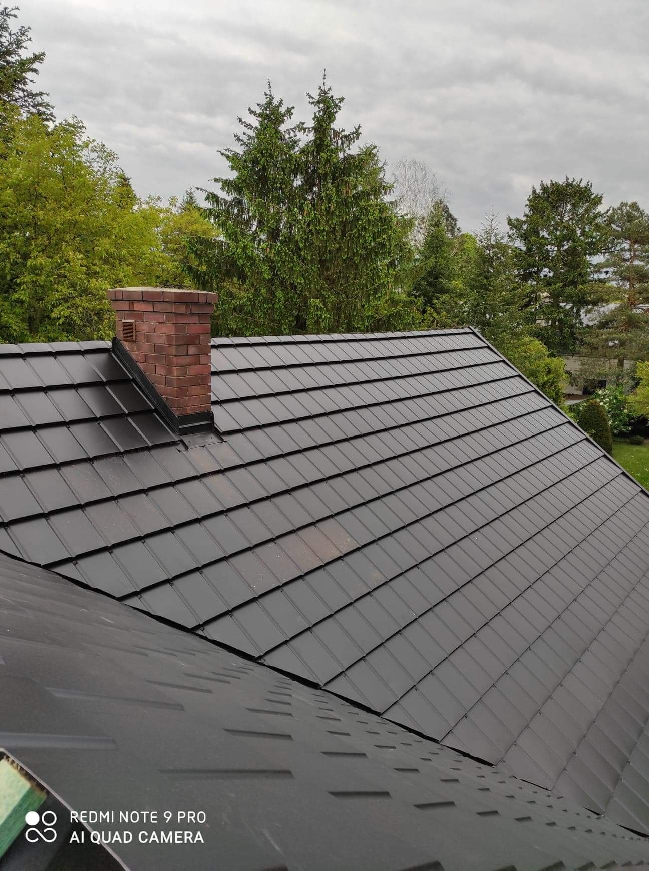 Nowy, czarny dach z metalowej blachy na tle zielonych drzew i pochmurnego nieba, z widocznym ceglanym kominem
