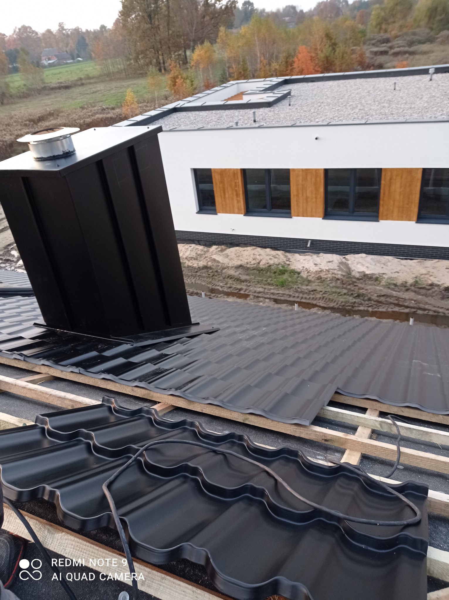 Montaż czarnej blachodachówki na drewnianej konstrukcji dachu, widoczny komin i fragment elewacji budynku w tle.