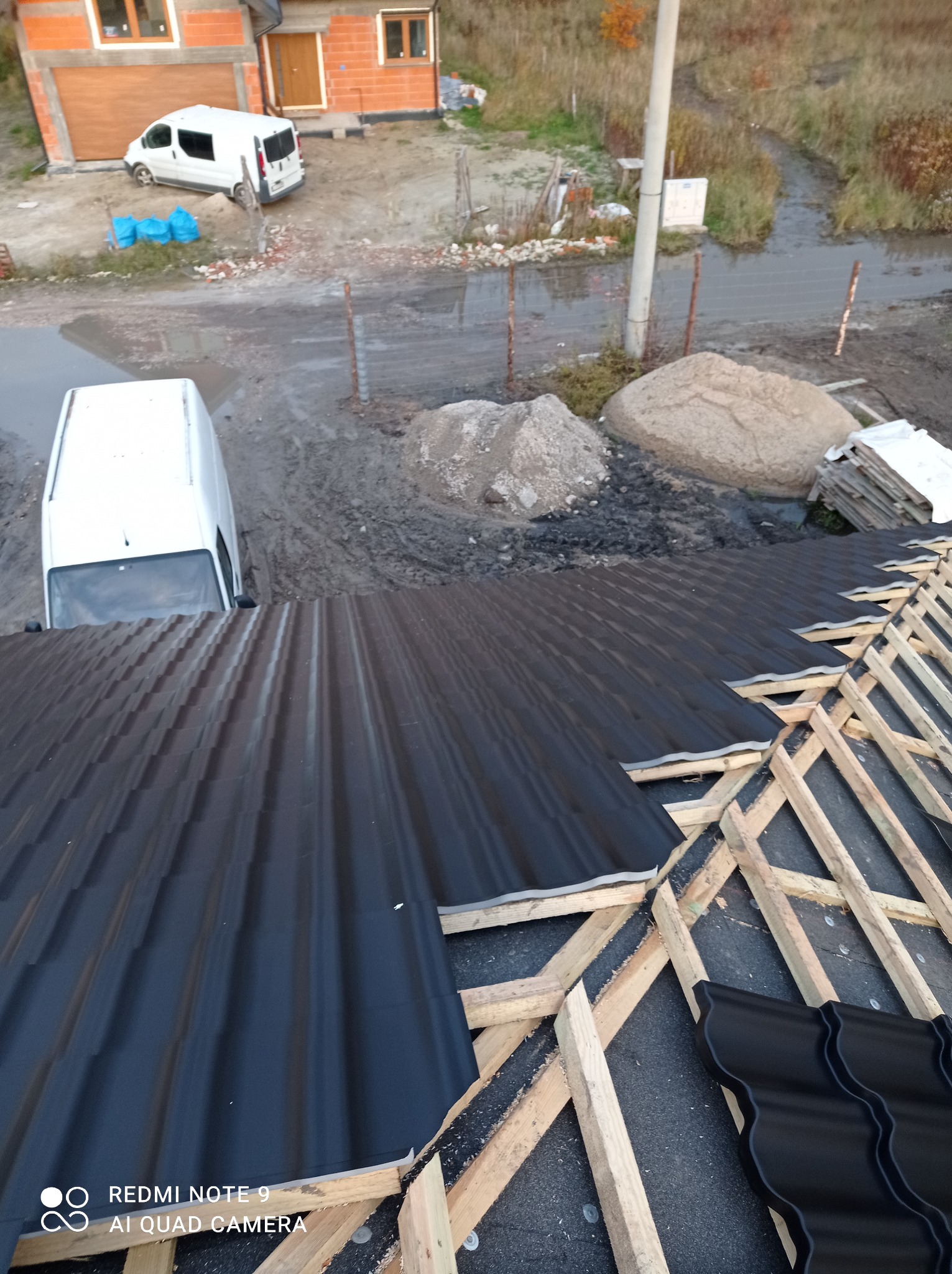 Montaż czarnej blachy na drewnianej konstrukcji dachu, widok z góry na częściowo pokryty dach, w tle dom w trakcie budowy i dwa białe busy.