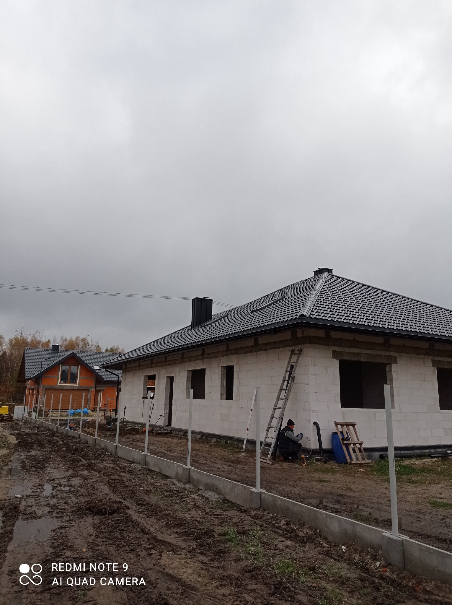 Dom w trakcie budowy z ciemnoszarym dachem z blachy, pracownik przy betonowym fundamencie ogrodzenia, pochmurne niebo.