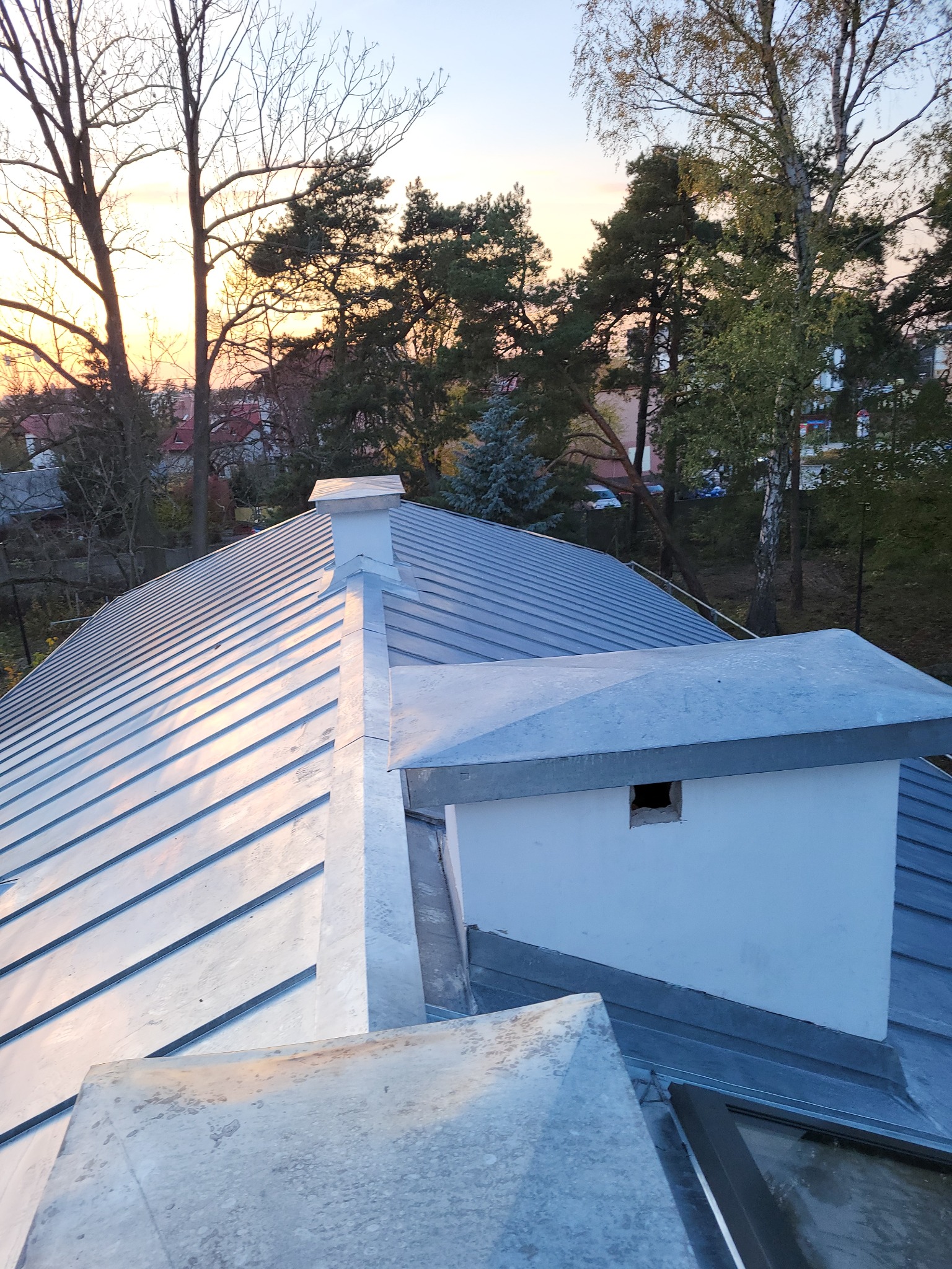 Metalowa powierzchnia dachu z kominem i elementami wentylacyjnymi, widok z góry na tle drzew i zachodzącego słońca.