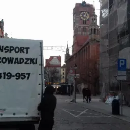 Plandeka samochodu dostawczego z napisem 'TRANSPORT PRZEPROWADZKI' i numerem telefonu na tle ulicy Starego Miasta z widoczną wieżą zegarową w Gdańsku.