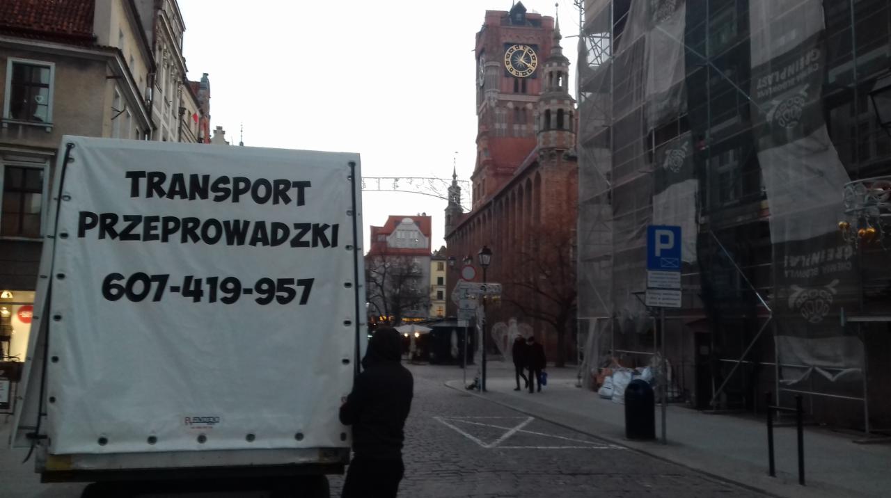 Plandeka samochodu dostawczego z napisem 'TRANSPORT PRZEPROWADZKI' i numerem telefonu na tle ulicy Starego Miasta z widoczną wieżą zegarową w Gdańsku.