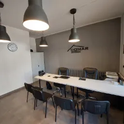 Nowoczesne biuro agencji nieruchomości z białym stołem konferencyjnym, czarnymi krzesłami, logo firmy DOMAR na szarej ścianie i trzema lampami wiszącymi w stylu industrialnym.