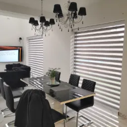 Jasne wnętrze jadalni z czarnymi meblami i poziomymi przesłonami okiennymi, widoczne lampy sufitowe z abażurami i telewizor w tle.