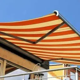 Pomarańczowo-biała, rozkładana markiza w paski nad balkonem z metalową balustradą i kwiatami w doniczce, widoczna na tle błękitnego nieba.