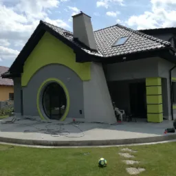 Nowoczesny dom jednorodzinny z elementami w kolorze limonkowym, okrągłym oknem i betonowym tarasem w trakcie wykończenia, widoczne przewody i narzędzia.