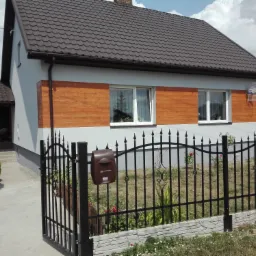 Dom jednorodzinny z szarą elewacją i panelami imitującymi drewno, widoczny zza czarnego, kutego ogrodzenia z brązową skrzynką pocztową Poczty Polskiej.