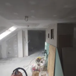 Poddasze w trakcie wykańczania, widoczne szpachlowane ściany, okno dachowe, narzędzia i materiały budowlane, w tym wiadro z farbą i szlifierka. Prace w toku, surowy stan.