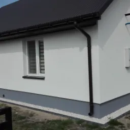 Świeżo pomalowany na biało dom jednorodzinny z szarą podmurówką i ciemnym dachem z blachy trapezowej, widoczne rynny i okna z roletami dzień-noc.