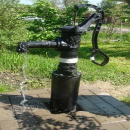 Żeliwna pompa ręczna z wylewającą się wodą, umieszczona na brukowanym podeście w ogrodzie.