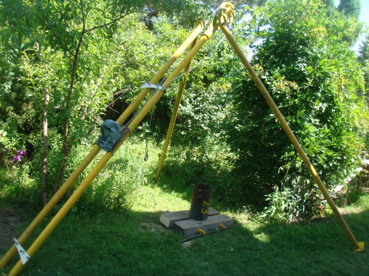 Żółty trójnóg z wciągarką ustawiony na trawie, obok metalowa rura z numerem 23, w tle zieleń ogrodu.