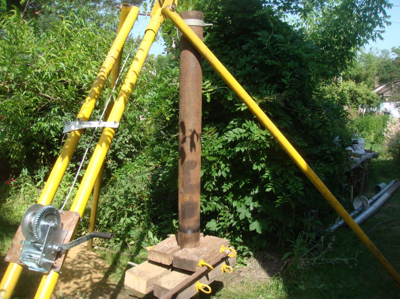 Żółty statyw wiertniczy z wciągarką linową i rurą wiertniczą na drewnianej podstawie, otoczony zieloną roślinnością.