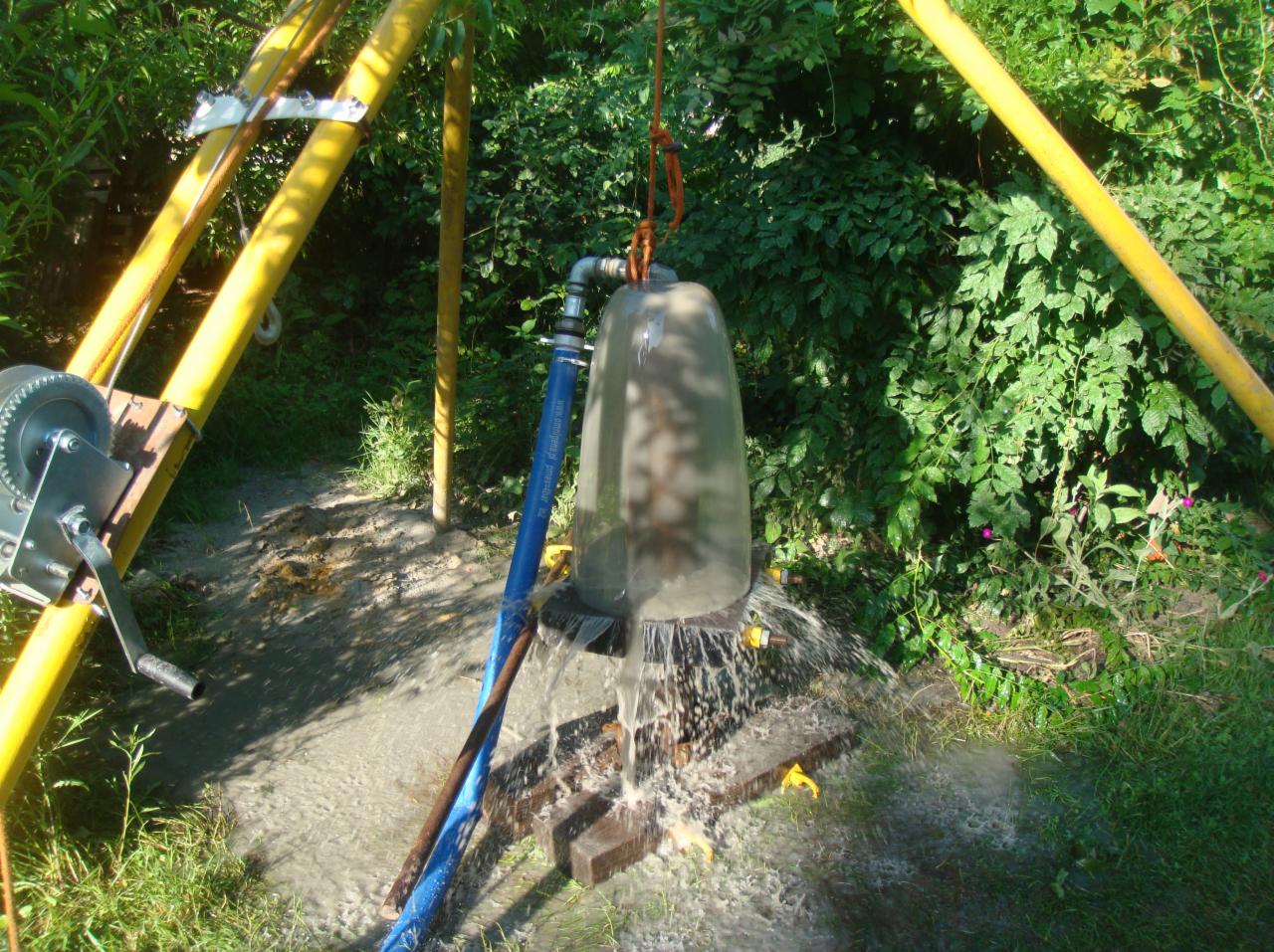 Testowanie wydajności studni głębinowej z użyciem pompy i przezroczystego separatora wody, woda wylewająca się na zewnątrz, widoczna konstrukcja wiertnicza z żółtych rur i lina mocująca.