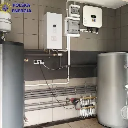 Instalacja grzewcza w pomieszczeniu technicznym z szarymi płytkami, widoczne dwa zbiorniki, biały bojler, rozdzielacz, skrzynki elektryczne i falownik. Na zbiorniku widoczne logo 'Polska Energia'.