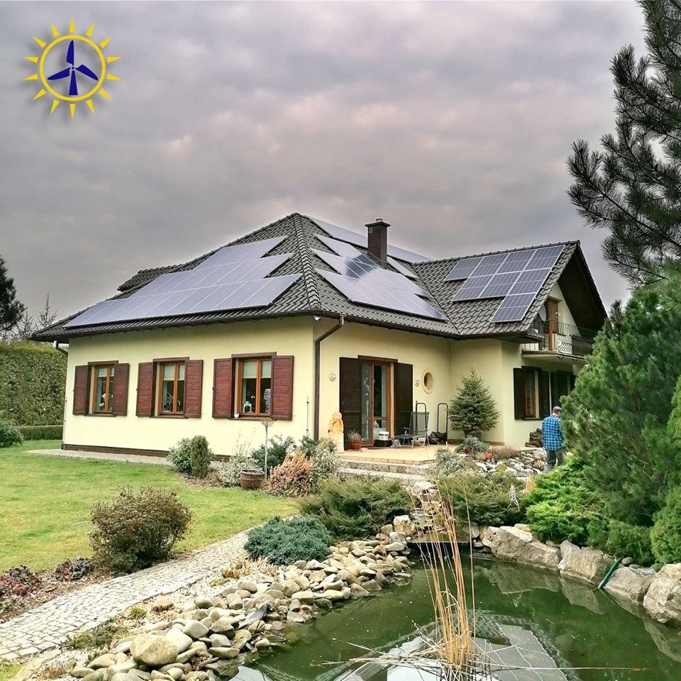 Dom jednorodzinny z panelami słonecznymi na dachu, widok z ogrodu z oczkiem wodnym i ścieżką z kostki brukowej, pochmurne niebo.