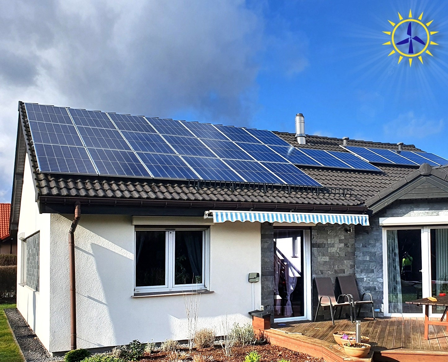 Dom jednorodzinny z panelami słonecznymi na dachu, widoczne także taras z meblami ogrodowymi i zadbany ogródek.