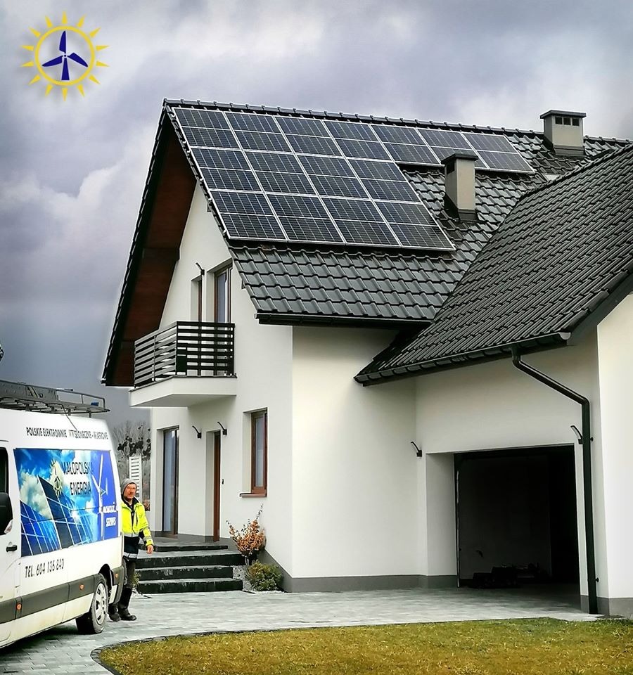 Biały dom jednorodzinny z zamontowanymi panelami słonecznymi na dachu, obok zaparkowany bus firmy z logo związanym z energią odnawialną, pochmurne niebo w tle.