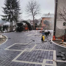 Ułożona kostka brukowa z jasnymi liniami dekoracyjnymi na posesji w zimowej scenerii, widoczny wózek z narzędziami i myjka ciśnieniowa, częściowo zasypane śniegiem otoczenie domu.