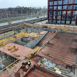 Rozległy plac budowy z widokiem z góry, ukazujący zbrojenie fundamentów pod budynek, z pracownikami w pomarańczowych kombinezonach i kaskach, z nowoczesnym budynkiem biurowym w tle i elementami...