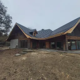 Nowoczesny dom z bali z czarnym dachem z blachy na rąbek stojący, widok z przodu na tle pochmurnego nieba, teren wokół domu w trakcie prac wykończeniowych.