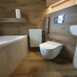 Nowoczesna łazienka z drewnopodobnymi płytkami na ścianach i podłodze, wisząca toaleta z chromowanym przyciskiem spłukiwania, biała wanna, grzejnik drabinkowy i uchwyt dla niepełnosprawnych.