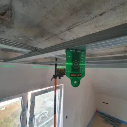 Montaż metalowego stelażu sufitu podwieszanego z płyt kartonowo-gipsowych, poziomowanego laserowym niwelatorem Stanley, widoczne profile i surowy betonowy strop.