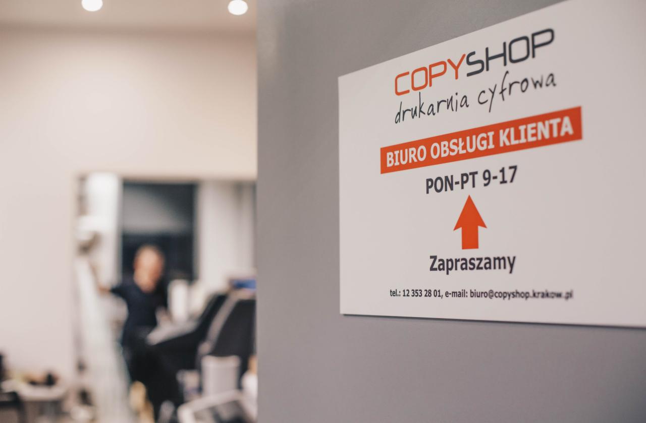 Tabliczka informacyjna drukarni cyfrowej COPYSHOP z godzinami otwarcia biura obsługi klienta, widoczna w tle postać w ruchu.