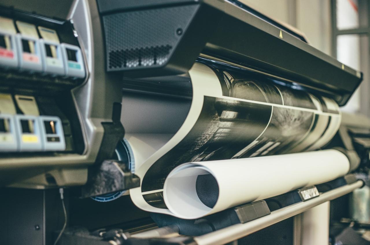 Drukarka wielkoformatowa w trakcie drukowania czarno-białego projektu graficznego, z widocznym podajnikiem papieru i kartridżami z tuszem.