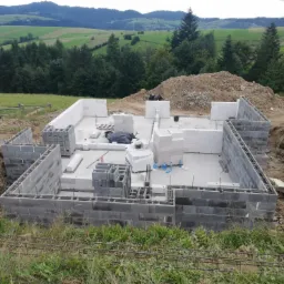 Wznoszenie ścian fundamentowych z bloczków betonowych na tle pagórkowatego krajobrazu. Widoczne zbrojenie pionowe i poziome, przygotowane pod wylewkę.