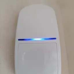 Biały czujnik ruchu alarmowego, zamontowany na jasnej ścianie, z widoczną niebieską diodą sygnalizującą aktywność.