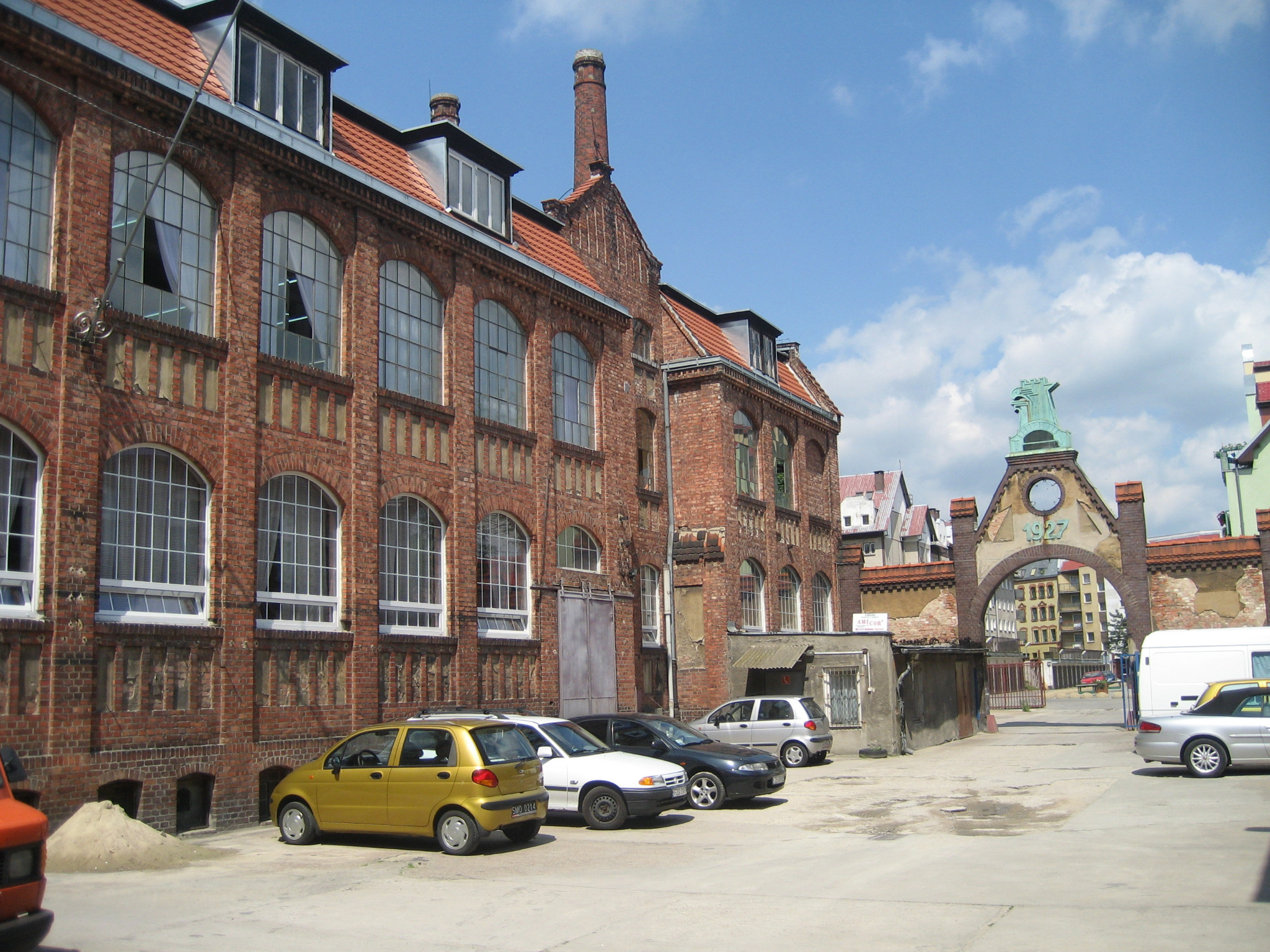 Ceglany budynek dawnej fabryki z wysokimi oknami i kominem, z zaparkowanymi samochodami i ozdobną bramą w tle, w słoneczny dzień.