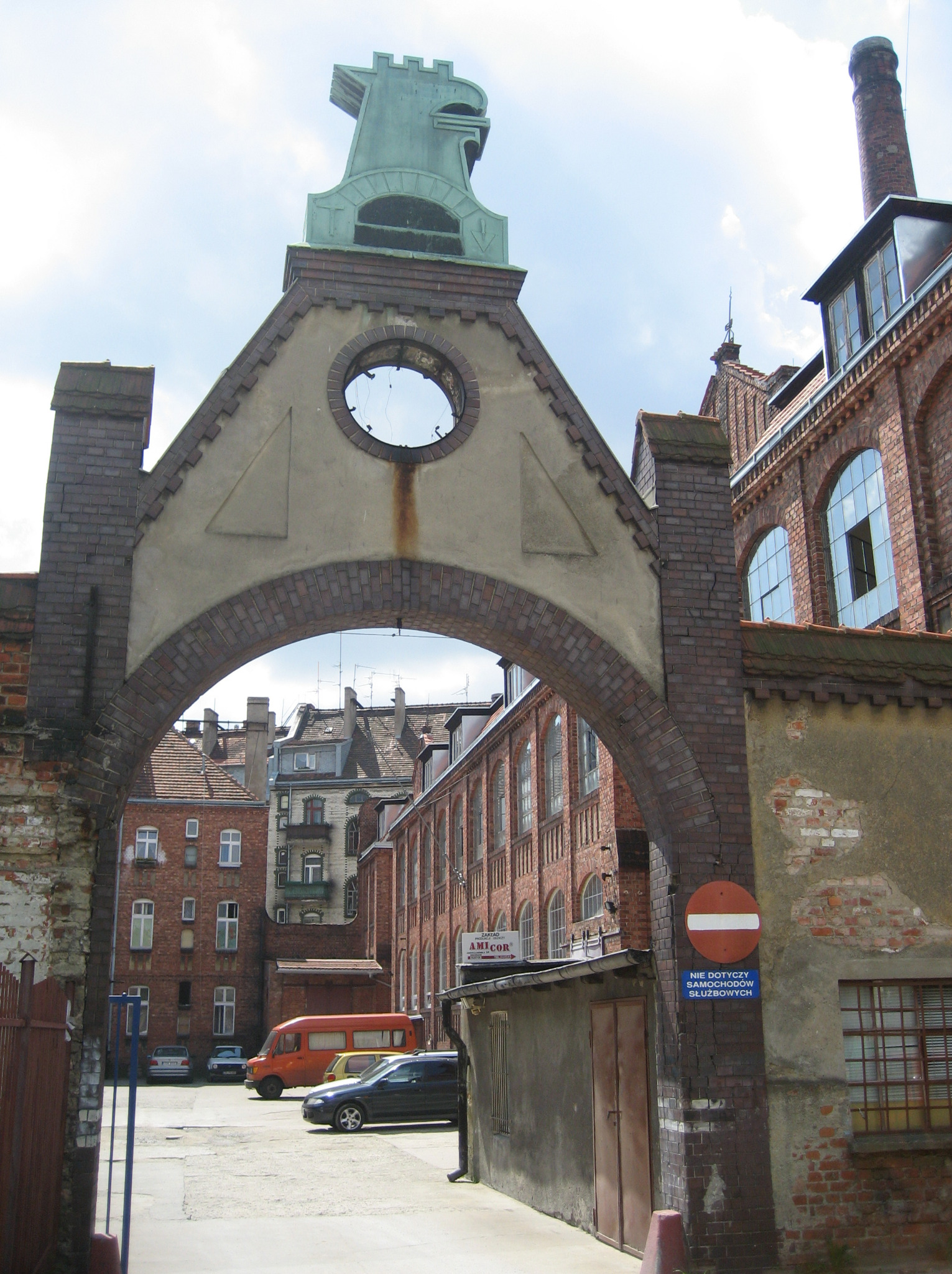 Fabryczny dziedziniec z ceglaną architekturą, w tle budynki z czerwonej cegły i zaparkowane samochody. Na pierwszym planie brama z okrągłym otworem.