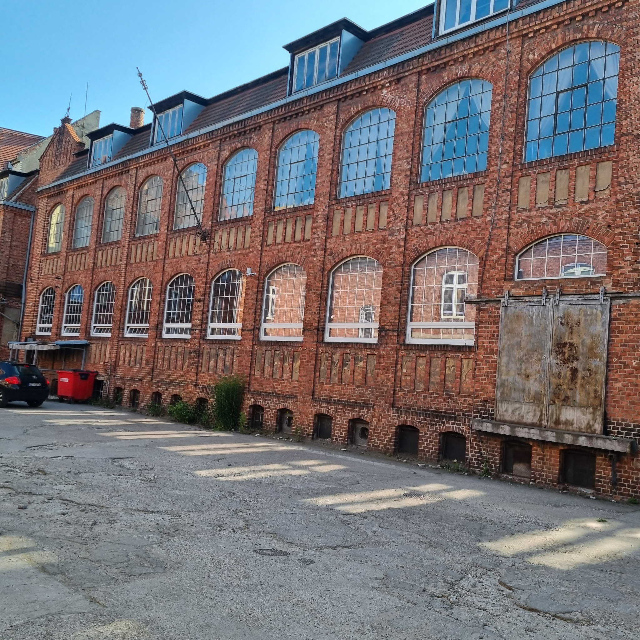 Fasada budynku z czerwonej cegły z dużymi oknami i metalowymi drzwiami. Widoczne elementy architektoniczne i otoczenie budynku.