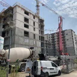 Wznoszenie wielopiętrowego budynku z betonu kom&oacute;rkowego; widoczny betonow&oacute;z, dźwig i pompa do betonu; pracownicy w kaskach na rusztowaniach.