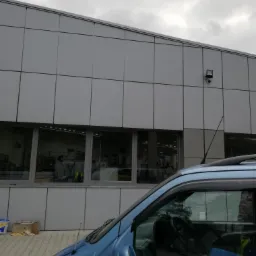 Szary budynek z panelami elewacyjnymi widziany z perspektywy boku zaparkowanego, niebieskiego samochodu dostawczego, z widocznym wnętrzem przez okna budynku.