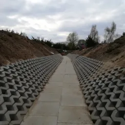 Ukończone koryto odwadniające z prefabrykatów betonowych, zabezpieczające skarpę przed erozją, widok z perspektywy pieszego w kierunku ujścia, pochmurne niebo.