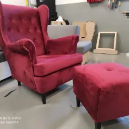 Czerwony, welurowy fotel typu uszak z podnóżkiem na tle warsztatu, widoczne narzędzia i inne meble w trakcie renowacji.