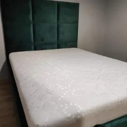 Łóżko z zielonym, tapicerowanym zagłówkiem w kształcie kwadratów i białym materacem w pokoju o szarych ścianach.