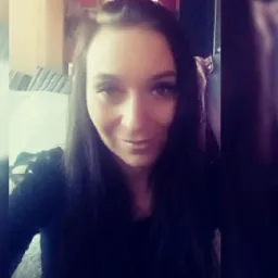 Portret uśmiechniętej kobiety z długimi, ciemnymi włosami, ubrana w czarną bluzkę, z widocznym fragmentem sofy w tle.